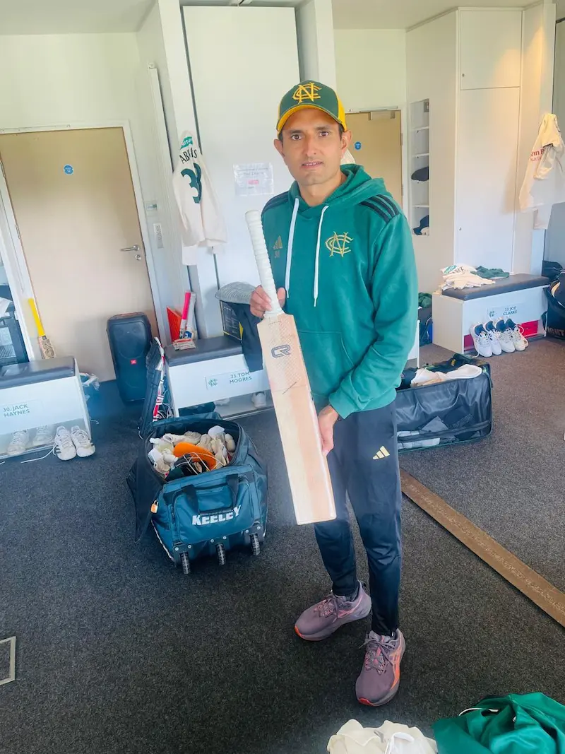Cricket player in team gear holding cr cricket bat in dressing room with kit bag and equipment